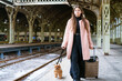 Travel concept. At station, young tourist with dog goes and drags suitcase with and looks for hotel on platform. Caucasian woman waiting for train and planning happy holiday vacation. In pink coat