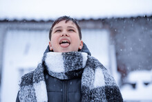 Portrait Of A Happy Young Man In A Winter Scarf. A Teenager, On Whom Snowflakes Fall In A Scarf In The December Season, Catches Snow With His Tongue