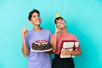 Young mixed race couple holding birthday cake and present isolated on blue background pointing with the index finger a great idea