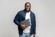 © Vadim Pastuh - Smiling young African-American male entrepreneur stands in studio isolated on gray and carrying trendy laptop. Cheerful black guy in glasses and casual shirt looks at camera, holding closed laptop