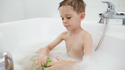 Wall Mural - Cute boy sitting in bath with soap foam and playing toy boats and ships. Concept of family time, children development and fun at home