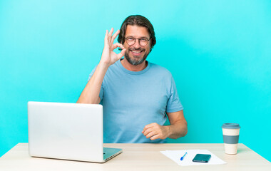 Senior dutch man in a table with a laptop isolated on blue background showing ok sign with fingers