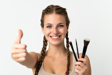 Wall Mural - Young caucasian woman isolated on white background holding makeup brush and with thumb up