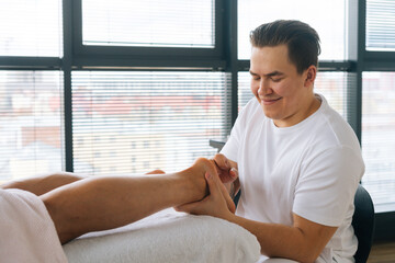 Wall Mural - Close-up of cheerful professional male masseur with strong hands massaging feet heels of muscular athlete man after physical sports workout, lying on stomach at massage table in salon by window.