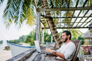 Technology and travel. Working outdoors. Freelance concept. Bearded young man using laptop in cafe on tropical beach.