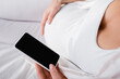 © LIGHTFIELD STUDIOS - cropped view of pregnant woman holding smartphone with blank screen.