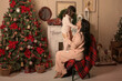 © Тамара Киреева - a beautiful brunette girl with long hair in pink pajamas is sitting on a chair covered with a plaid plaid by the Christmas tree fireplace holding a little daughter