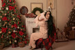 © Тамара Киреева - a beautiful brunette girl with long hair in pink pajamas is sitting on a chair covered with a plaid plaid by the Christmas tree fireplace holding a little daughter
