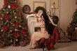 © Тамара Киреева - a beautiful brunette girl with long hair in pink pajamas is sitting on a chair covered with a plaid plaid by the Christmas tree fireplace holding a little daughter