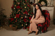 © Тамара Киреева - a beautiful brunette girl with long hair in pink pajamas is sitting on a chair covered with a plaid plaid by the Christmas tree fireplace