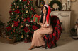 © Тамара Киреева - a beautiful brunette girl with long hair in a Santa Claus hat in pink pajamas is sitting on a chair covered with a plaid plaid by the Christmas tree fireplace in her hands a gift box