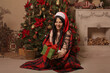 © Тамара Киреева - a brunette girl in a Santa Claus hat in pink pajamas is sitting covered with a plaid plaid by the fireplace in her hands a gift box Christmas tree new year