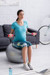 © LIGHTFIELD STUDIOS - pregnant sportswoman exercising on fitness ball with dumbbells near sports bottle and laptop.