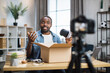 © sofiko14 - African american man in casual wear opening parcel box and recording video at home. Male influencer sitting at table with VR glasses in hands and talking on camera.