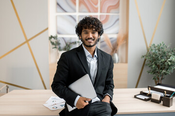 Portrait of a confident successful Indian businessman, manager, IT specialist, stock trader, in formal suit, stands near his desk in the office holds a laptop,looks at the camera with a friendly smile