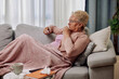© Space_Cat - Sick elderly woman checking her temperature with thermometer lying on sofa caughing suffering from seasonal flu or cold. Ill senior feel unhealthy with influenza at home