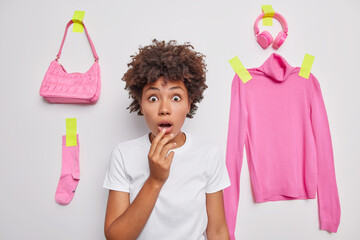 Wall Mural - Stunned curly haired woman sees something incredible in front gasps from wonder dressed in casual basic t shirt poses against white background with plastered clothes around being speechless.