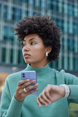 Wall Mural - Vertical shot of thoughtful curly haired young woman waits for someone checks time on watch holds mobile phone sends sms to friend wears casual jumper poses outside against blurred background