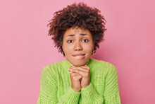 Hopeful Worried Curly Haired Young Woman Keeps Hands Under Chin Bites Lips Looks With Pleading Expression At Camera Says Please Or Sorry Wears Warm Knitted Sweater Isolated Over Pink Background.
