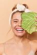© WHstudio Leushin N - Positive woman with healthy glowing skin uses natural cosmetics covers face with leaf of cabbage wears headband stands bare shoulders feels very happy to undergo facial treatments stands indoor