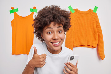 Wall Mural - I like it. Pleased curly haired woman keeps thumb up holds mobile phone recommends new application smiles gladfully poses around clothes items isolated over white background makes shopping online