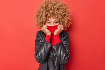 Waist up shot of happy curly haired young woman covers mouth with collar of turtleneck wears leather jacket expresses positive emotions stands against red background hides emotions foolishes around