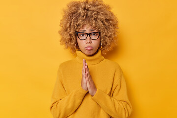 Wall Mural - Troubled upset woman keeps palms pressed together feells unhappy looks with pleading expression wears spectacles and casual jumper isolated over yellow background. Please help me one more time