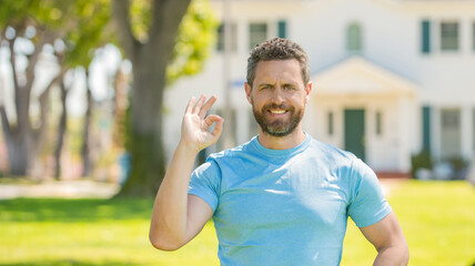 happy mature man realtor standing outdoor at house showing ok gesture, buying house