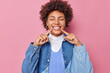 Positive young curly woman keeps eyes closed points with fingers at toothy smile shows perfect teeth wears denim shirt expresses happy emotions isolated over pink background. Happiness concept