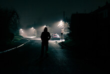 A Moody Concept Of A Scary Hooded Figure. Standing In The Middle Of The Road In A Quiet Suburban Street. On A Foggy Winters Night