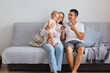 © sementsova321 - Indoor shot of attractive happy young couple wearing casual style clothing playing with their little daughter, sitting on sofa in light living room with infant baby.