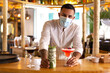 © Alfredo López - Elegant young Latin American bartender serving a cocktail at the bar of a modern cocktail bar. He wears a surgical mask while working.