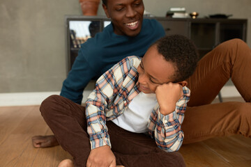 Wall Mural - Funny indoor crop shot of dad and son resting on floor in living-room against home interior background, laughing, joking, having fun, dark-skinned boy in lotus posture supporting his head in his hand