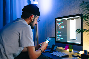 Young man mobile developer writes program code on a computer and check app in smartphone, programmer work in home office.