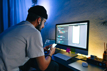 Young man mobile developer writes program code on a computer and check app in smartphone, programmer work in home office.