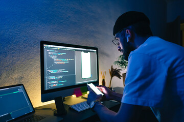 Young man mobile developer writes program code on a computer and check app in smartphone, programmer work in home office.