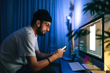 Young man mobile developer writes program code on a computer and check app in smartphone, programmer work in home office.