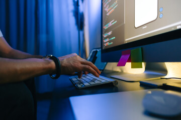 Mobile developer writes program code on a computer, typing on keyboard, programmer work in home office.