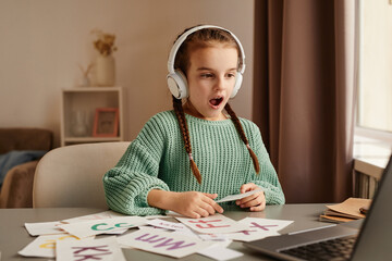 Little girl in wireless headphones sitting at the table and learning to pronounce new english words during online lesson on laptop