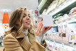 © Анна Демидова - Young woman chooses sweet cake in the supermarket. A beautiful blonde woman in a beige coat. Close-up.