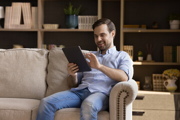 Canvas Print - Happy young man holding digital device, resting on couch at home, using online virtual app on tablet, browsing internet, chatting, reading book, watching multimedia content, laughing