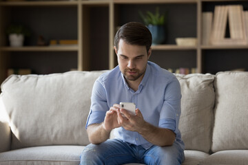 Wall Mural - focused millennial guy un casual using cellphone on couch at home, typing, chatting online, reading 