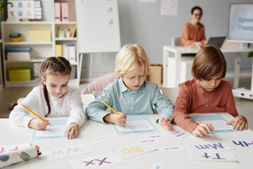 Group of school children sitting at the table and making notes on papers during lesson at school with teacher in the background