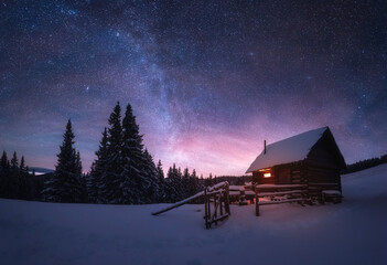 Fantastic winter landscape with wooden house in snowy mountains. Starry sky with Milky Way and snow covered hut. Christmas holiday and winter vacations concept