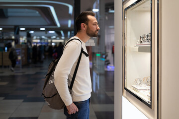Photo of an adult doubtful thoughtful thinking handsome brunette man in a white sweatshirt and with a backpack chooses a stylish wrist watch in a store in a shopping center