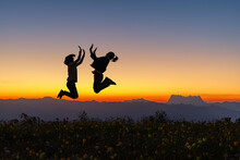 Silhouette Of Happy Jumping Teenager Girlfriend On Flowers Mountain At Colorful Sunset Sky Background.