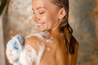 Beautiful young woman applying shower gel using loofah sponge while taking shower. Spa and body care concept
