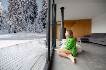 Woman sitting with dog near fireplace and panoramic window at modern living room with stunning view on snowy mountains. Concept of rest in houses or cabins on nature. Idea of escape from everyday life