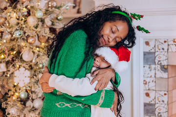 African American woman with curly hair and brunette daughter pose hugging and wearing Christmas hats against tree decorations