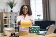 © sofiko14 - Smiling african woman in casual wear sitting at desk and holding colorful gift boxes in hands. Famous female influencer opening festive presents made by worldwide companies.